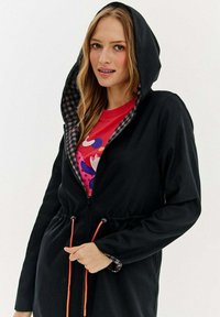 Sieviete, kura valkā melnu kapuci jaku ar oranžiem savilcējiem virs krāsainas sarkanās krekla, tur jaku ar vienu roku aiz savilcējiem.