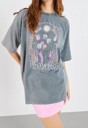 Nainen, joka pukeutuu ylisuurikokoiseen harmaaseen T-paitaan, jossa vaaleanpunainen ja valkoinen tulppaanikuva, aurinko, rauhanmerkit ja teksti "LOVE YOURSELF" ja "FLOWERS."