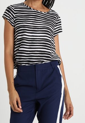 Vrouw draagt een zwart-wit gestreept kortarmig topje, ingestopt in marineblauwe broek met een witte zijstreep, staat met ontspannen handen.