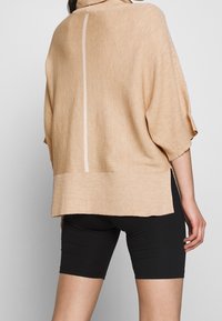 Maglione beige oversize con texture a costine, maniche corte, collo alto e una linea bianca verticale sul retro, abbinato a pantaloni shorts neri.