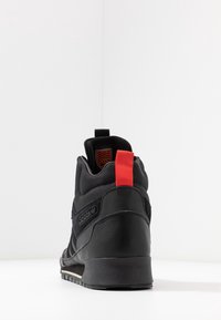 Baskets montantes noires avec une tige synthétique texturée, un languette rouge au talon et une semelle en caoutchouc avec un motif de bande de roulement visible.