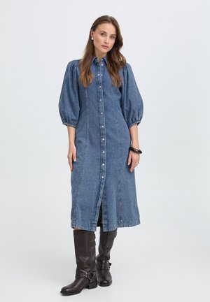 Vrouw staat en draagt een blauwe denim midi-jurk met knopen, pofmouwen en zwarte kniehoge leren laarzen tegen een effen lichte achtergrond.