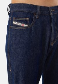 Blå denimjeans med rak skärning, orange sömmar och en logotags i bakfickan. Tyget har en klassisk textur.