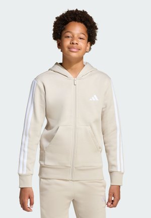 Ragazzo adolescente in piedi, indossa una felpa con cappuccio beige Adidas e pantaloni abbinati con strisce bianche lungo le maniche e logo sul petto e sui pantaloni.