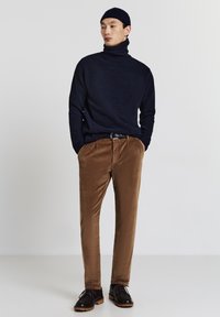 Marineblauer Rollkragenpullover, braune Cordhose und dunkle Schuhe. Das Outfit zeichnet sich durch einen strukturierten Stoff und einen entspannten Schnitt aus.