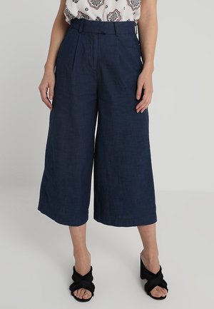 Frau trägt marineblaue, weite, knöchellange Hose mit hohem Bund, kombiniert mit schwarzen Sandalen mit Absatz und gekreuzten Riemen.