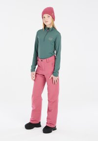 Dlouhý zelený termo top s límcem na zip, zkombinovaný s růžovými voděodolnými lyžařskými kalhotami. Černé sněhule dokreslují celkový vzhled.