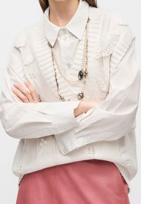 Personne portant un gilet en tricot blanc texturé sur une chemise blanche à col, les bras croisés et un long collier de perles, associé à une jupe rose.