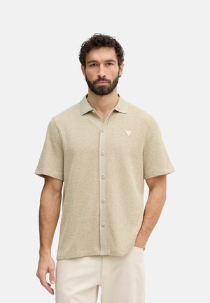 Hombre con barba que lleva una camisa de punto beige de manga corta con botones y cuello, de pie con una mano en el bolsillo contra un fondo liso.