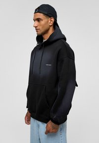 Zwarte gradient hoodie met een voorzak, een trekkoordkap en logotekst. Heeft een losse pasvorm en een zachte textuur van de stof.