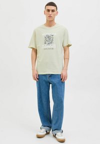 Lys grøn grafisk t-shirt med sort blomsterdesign, parret med løse blå jeans og hvide sneakers med brune accenter.