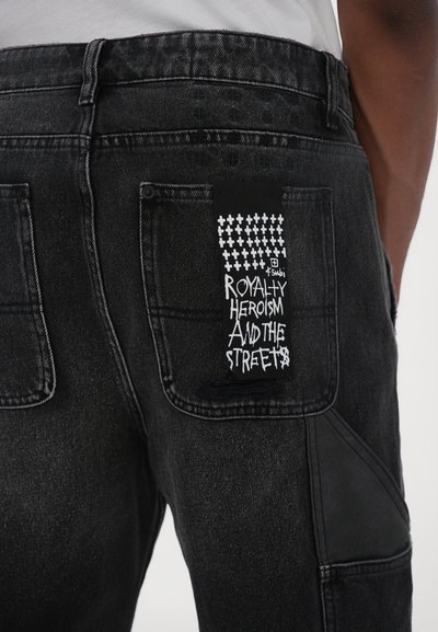 Jeans en denim noir avec une étiquette texturée sur la poche arrière, portant un texte graphique blanc indiquant "ROYALTY HEROISM AND THE STREETS". Accents en cuir noir.