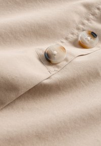 Beiger Stoff mit glatter Textur, der zwei runde, gesprenkelte Knöpfe mit hellem und dunklem Muster aufweist, die am Kleidungsstück angenäht sind.