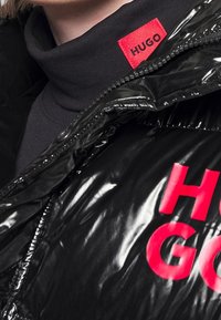 Czarna kurtka puchowa o błyszczącym wykończeniu, z odważnym czerwonym napisem "HUGO" oraz wysokim, czarnym kołnierzem wystającym z dekoltu.