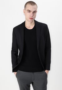 Zwarte op maat gemaakte blazer over een zwarte gebreide trui, gecombineerd met grijze broeken. Glad materiaal, gestructureerde pasvorm en minimalistisch ontwerp.