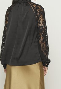 Blouse en satin noir avec manches longues en dentelle noire, dotée d'un col froncé et de poignets boutonnés, associée à une jupe en satin doré.