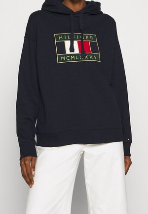 Personne portant un sweat à capuche noir avec un logo rectangulaire multicolore et un pantalon blanc, debout devant un fond uni.