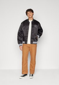 Chaqueta bomber de satén negra con puños a rayas blancas, logo en el lado izquierdo del pecho, llevada sobre una camisa clara y combinada con pantalones marrones y zapatillas negras.