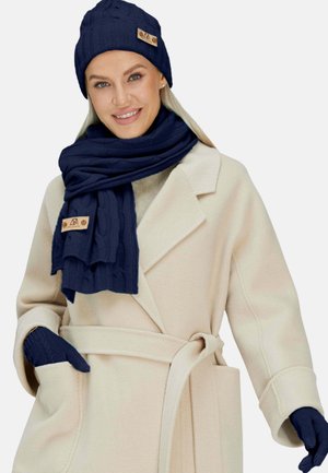 Berretto in maglia navy, sciarpa e guanti con motivi a treccia; dettaglio in pelle con logo; indossati con un cappotto avvolgente di un leggero beige.