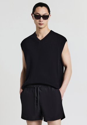 Black sleeveless sweater with a V-neck design, paired with black drawstring shorts. Sleek, smooth fabric texture with a relaxed fit.