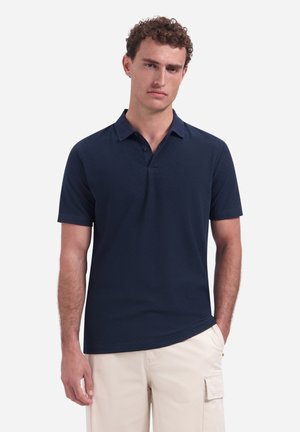 Man die een donker marineblauw poloshirt en lichtbeige cargobroek draagt, staand met één hand in de zak tegen een effen achtergrond.