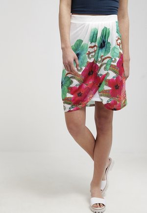 Femme portant une jupe portefeuille blanche à grand imprimé floral rouge et vert, associée à des sandales blanches et un haut sans manches bleu marine.