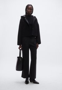 Veste noire courte avec capuche, pull noir côtelé, pantalon large noir et bottines noires. Le mannequin tient un sac fourre-tout noir.