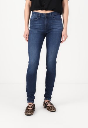 Jeans ajustados en denim azul oscuro, con cintura alta, cinco bolsillos y costuras sutiles. Combinados con una blusa negra y bailarinas con estampado.