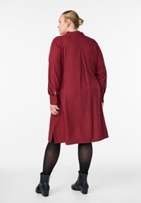 Červené pruhované šaty s dlhými rukávmi, manžetami na gombíky a bočnými roztrhnutiami, vyrobené z textúrovanej látky. Zadaj obsahujú vertikálny šev a golier.