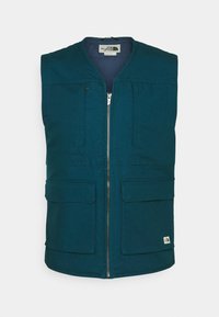 Petroliumsfärgad ärmlös väst med dragkedja fram, två stora framfickor och subtilt texturerad tyg. Har en etikett nära fållen.