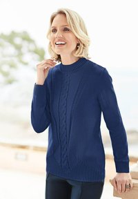 Blonde vrouw die glimlacht, gekleed in een blauwe kabelgebreide trui, staande in de buitenlucht met een vervaagde groene achtergrond en een lichte achtergrond.