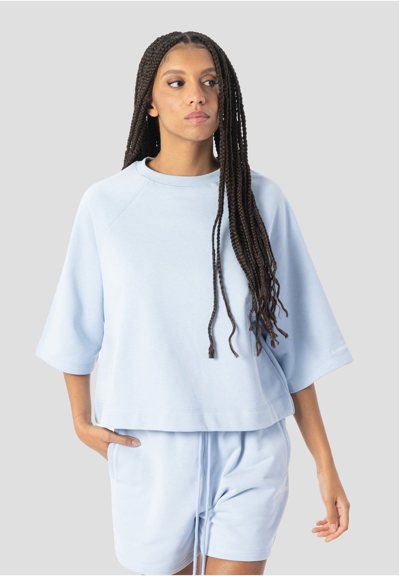Femme aux cheveux longs tressés portant un sweat-shirt ample à manches courtes bleu clair et un short assorti, debout avec une main dans la poche.