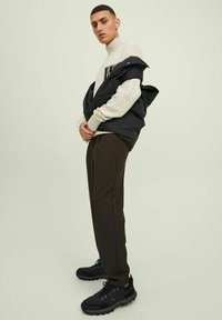 Chaqueta de plumas negra sobre un suéter crema con logo, pantalones marrones y zapatillas negras. La modelo está de perfil contra un fondo claro.