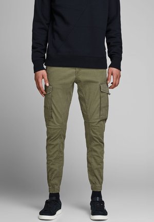 Hombre con pantalones jogger cargo de color verde oliva con bolsillos laterales, una sudadera negra, calcetines negros y zapatillas negras con suelas blancas.
