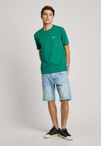 Camiseta de algodón verde con mangas cortas y un pequeño logotipo, combinada con pantalones cortos de mezclilla azul claro y zapatillas negras de corte bajo.