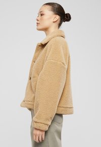 Giacca in fleece beige con colletto, taglio ampio e dettagli di cucitura visibili; presenta tasche grandi e una texture morbida.