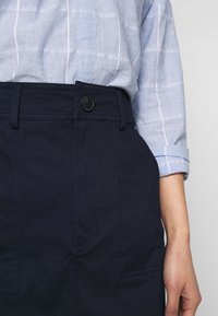 Marinblå högmidjade byxor med sidofickor, med knappstängning fram och rak bendesign. Skjortan har ett ljusblått randigt mönster.