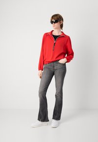 Rode zip-up jack met elastische manchetten, gedragen over een zwart topje, gecombineerd met grijze flared jeans en witte sneakers. Model draagt donkere zonnebril.