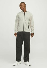 Veste zippée gris clair avec capuche et accents noirs, associée à un pantalon noir ample et des baskets blanches, sur un fond neutre.
