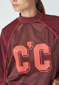 Maroon urheilu paita, jossa on verkkokudonta, korkea kaulus ja neonoranssi "C'C" logo. Ominaisuuksiin kuuluvat kontrastiväriset olkaakkentit.