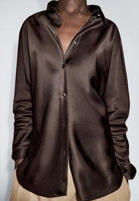 Braunes Satin-Hemd mit Knopfleiste und Kragen, ausgestattet mit langen Ärmeln und einer glatten, glänzenden Textur, kombiniert mit beigen Hosen.