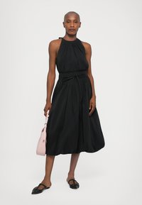 Rochie neagră cu gât halter, din material pliat, cu curea la talie și fustă amplă. Modelul ține în mână o poșetă roz și poartă pantofi negri cu talpă plată.