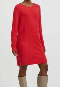 Robe en maille rouge avec un col rond, manches longues et poignets côtelés. Texture douce, coupe légèrement ample, longueur au-dessus des genoux.