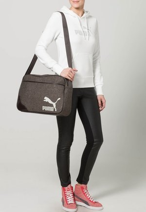 Persona con felpa bianca Puma con cappuccio e leggings neri che tiene una borsa a tracolla marrone Puma, indossando sneakers alte rosa Puma.