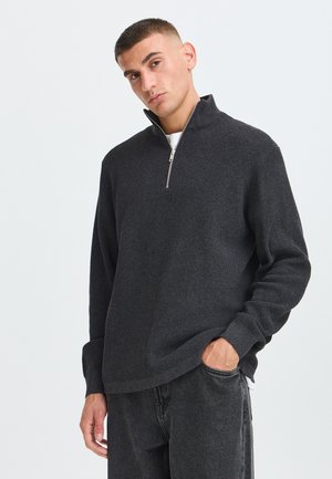 Ung mand iført en mørkegrå tekstureret halv lynlås sweater og sorte jeans, stående med den ene hånd i lommen mod en ensfarvet lys baggrund.