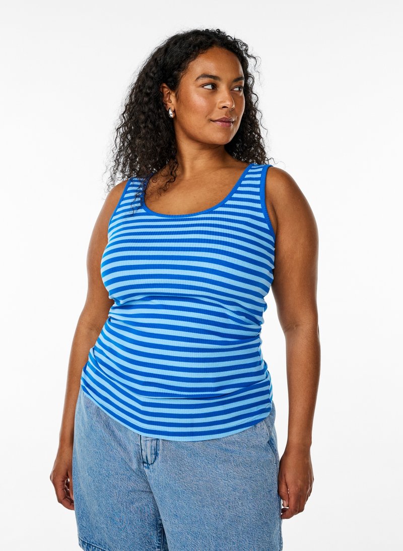 Kvinde med krøllet hår iført blå stribet tanktop og lyseblå denimshorts, der kigger til siden mod en hvid baggrund.