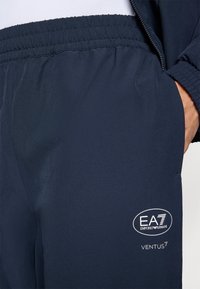 Temno modri atletski hlače z elastičnim pasom, ki vključujejo belo logotipno oznako, gladko teksturo in elegantno silhueto.