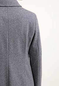 Närbild av en person som bär en grå ullkappa, som visar detaljerna på ryggen och axelsömmen mot en enkel ljus bakgrund.