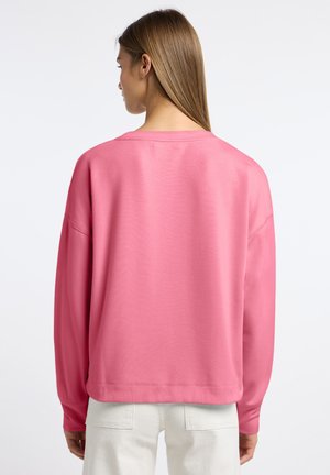 Kobieta o prostych, jasnobrązowych włosach nosi luźną różową bluzkę z długim rękawem i białe spodnie, pokazana od tyłu na białym tle.