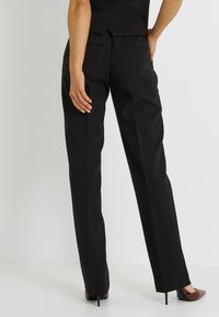 Pantalones negros a medida con un corte recto, con una textura lisa y dos bolsillos traseros, combinados con tacones de punta afilada.